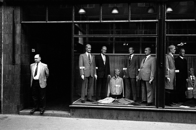 AKG1008103.webp Homme Devant Une Vitrine De Mannequins En Costumes – Image 1