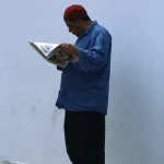 Homme lisant un journal adossé à un mur blanc.