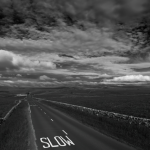 Route de campagne avec inscription "SLOW" sous un ciel nuageux en noir et blanc