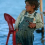 Enfant en salopette près d’une chaise rouge au bord de l'eau