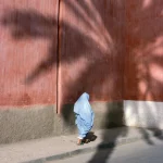 Femme en bleu marchant le long d'un mur orné de palmiers en ombre