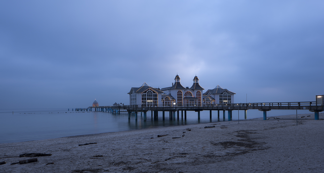 AKG2425876.webp Ponton sur la plage à l'aube avec bâtiments éclairés – Image 1