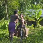 Enfant sur un vélo accompagné d'un adulte dans la nature tropicale.
