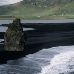 Plage de sable noir avec rocher en premier plan et montagnes en arrière-plan