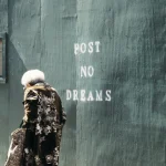 Personne âgée marchant devant un mur avec un graffiti "Post No Dreams"