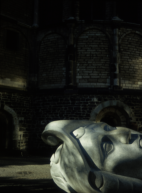 AKG5616063.webp Tête sculptée en pierre devant un bâtiment en briques – Image 1