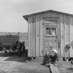 Homme assis devant une cabane avec une voiture ancienne à côté