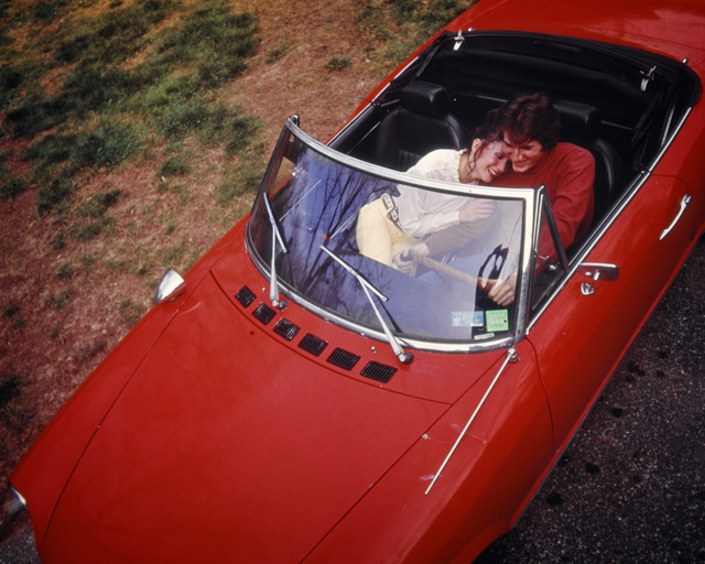 AKG7727272.webp Couple souriant dans une voiture décapotable rouge – Image 1