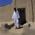 Homme en robe traditionnelle devant une maison en terre cuite
